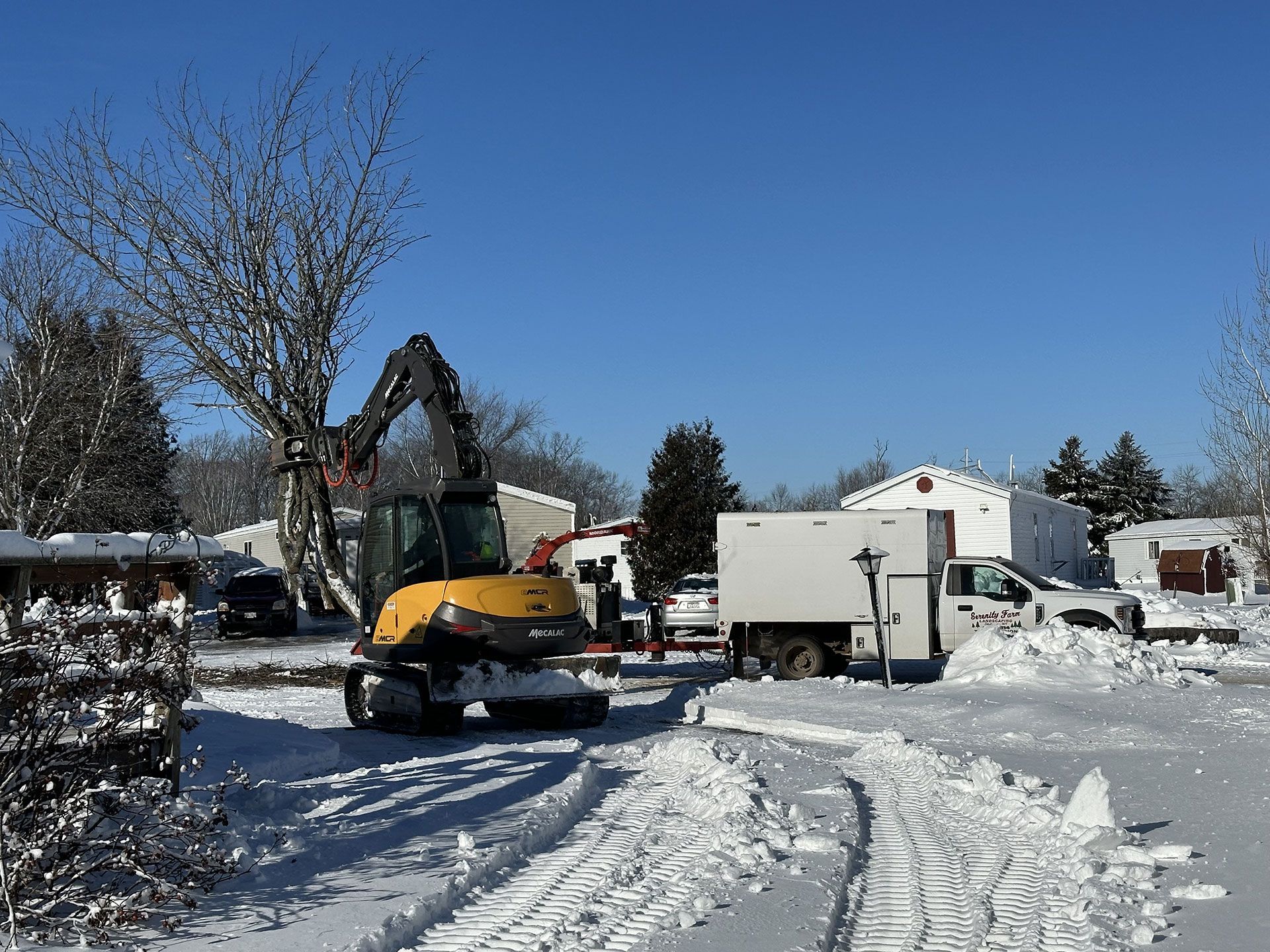A yellow construction excavator with a grapple attachment parked in a snow-covered yard near a white utility truck.