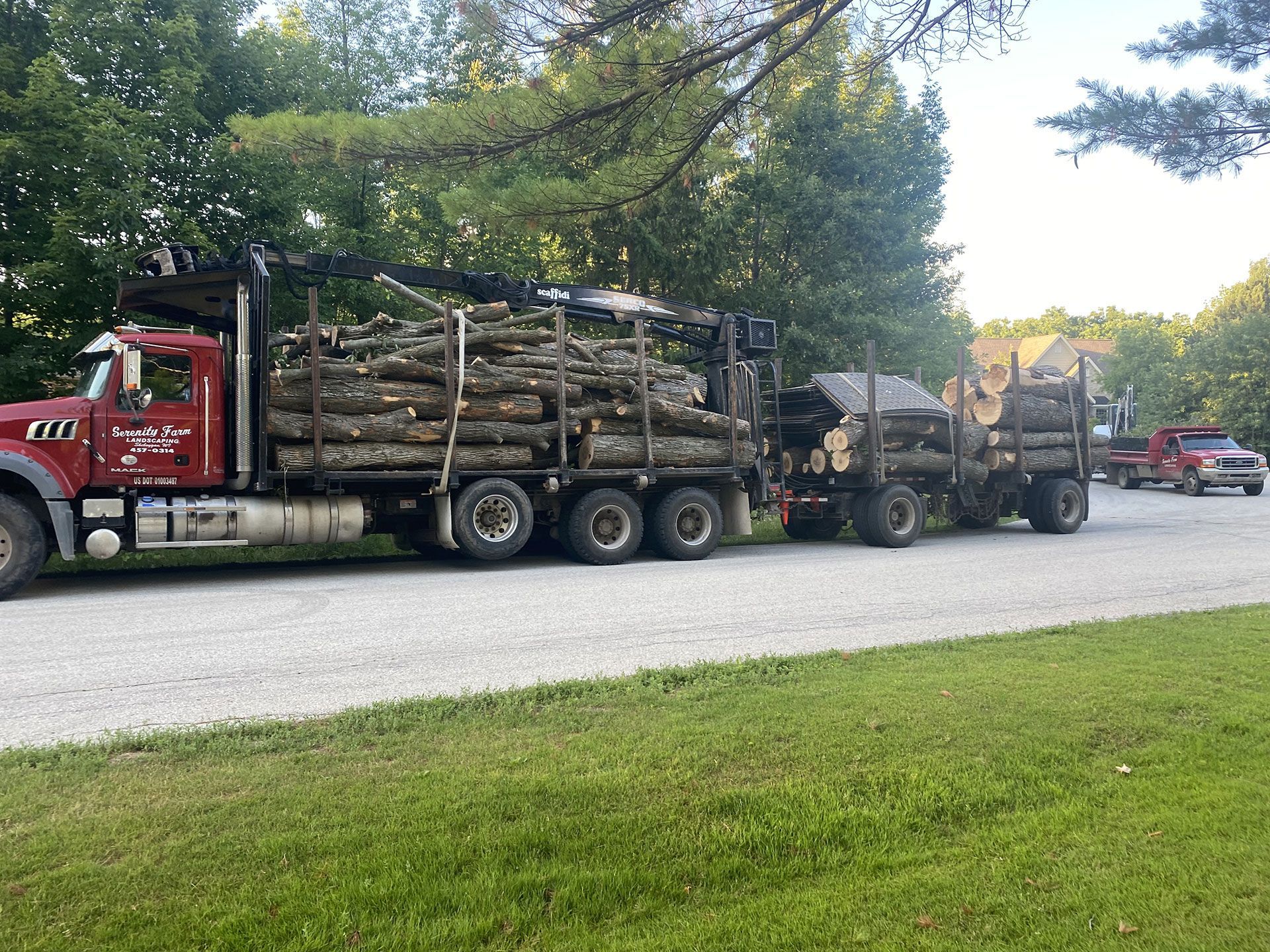 A red logging truck with a trailer loaded with timber is parked on a gravel road near trees.