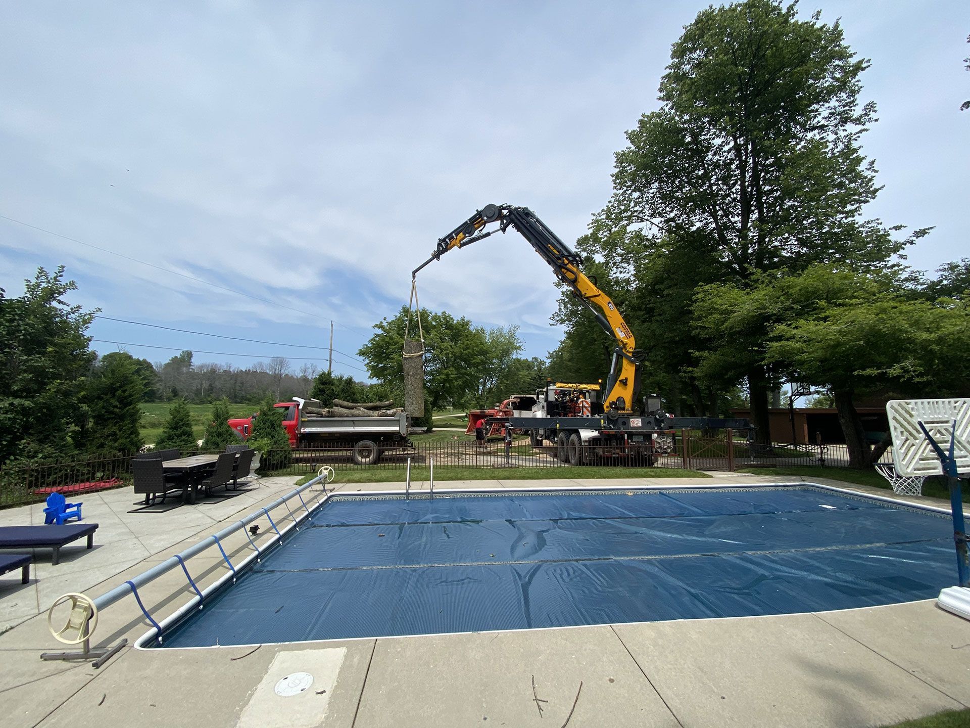 A crane lifts a large, vertical object near a swimming pool covered by a blue solar tarp on a sunny day.