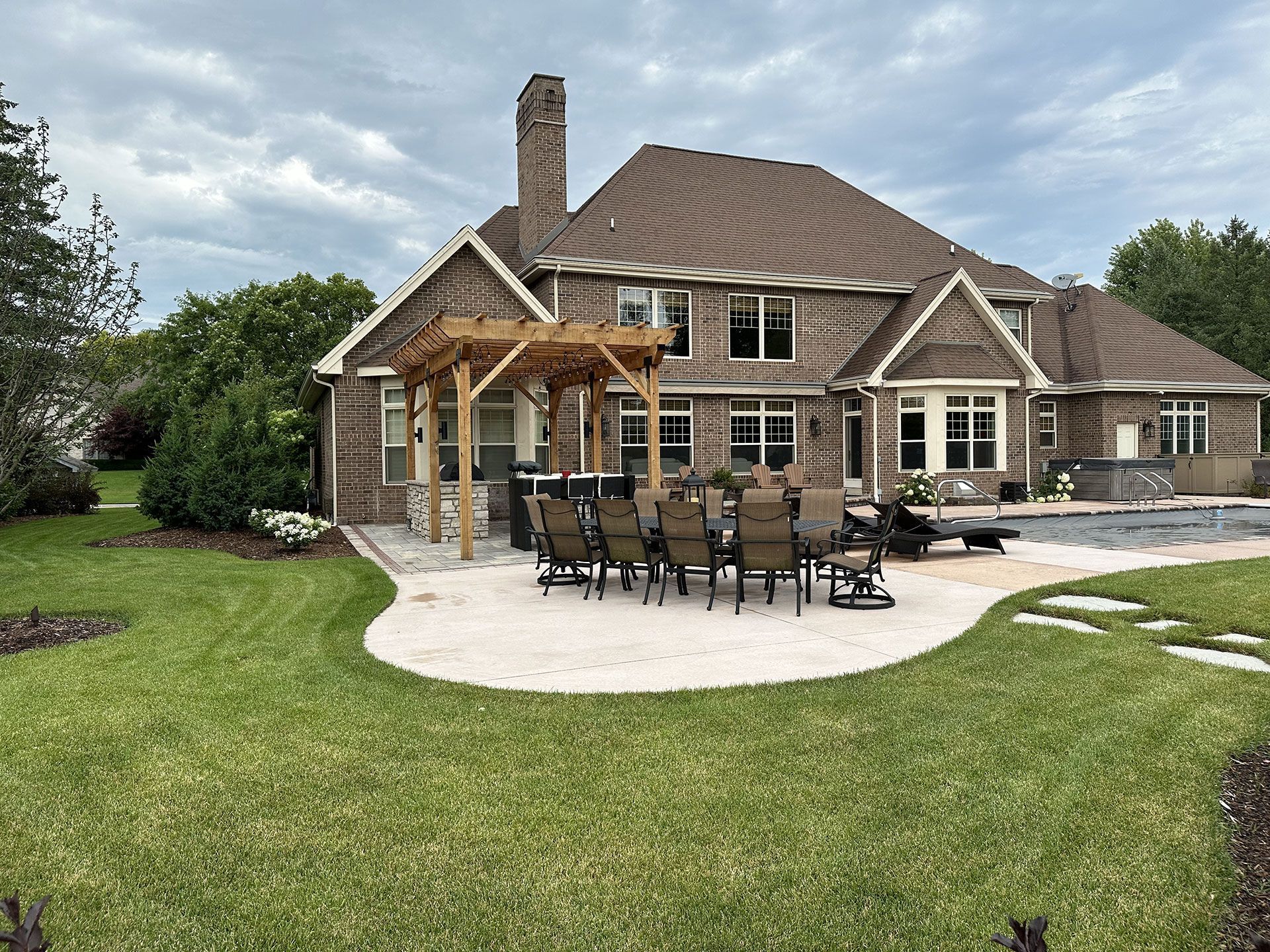 A stone patio with a wooden pergola, dining set, and pool behind a large brick house under a cloudy sky.