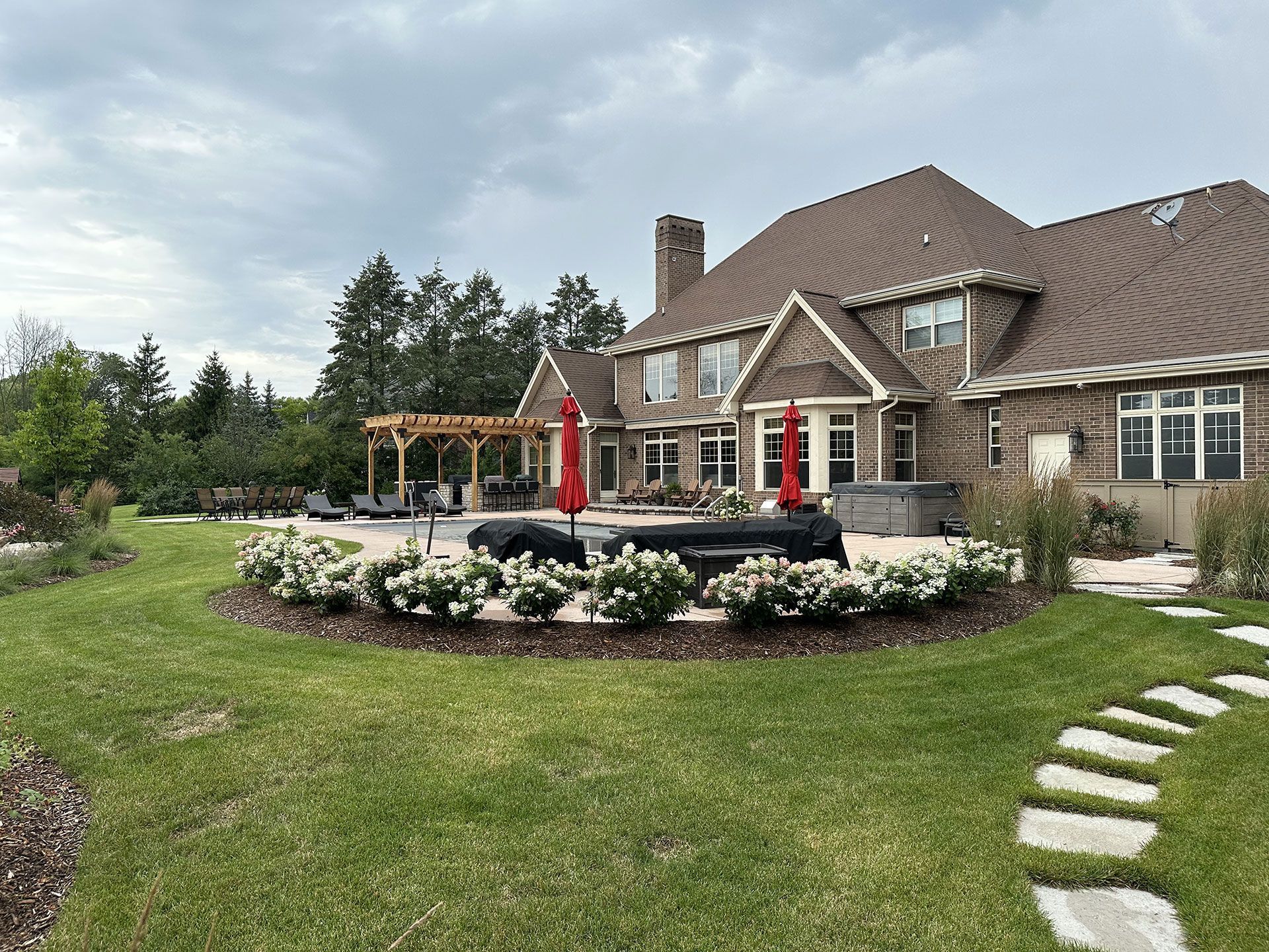 A backyard view of a large brick house with a patio, swimming pool, pergola, and a curved garden bed with white flowers.