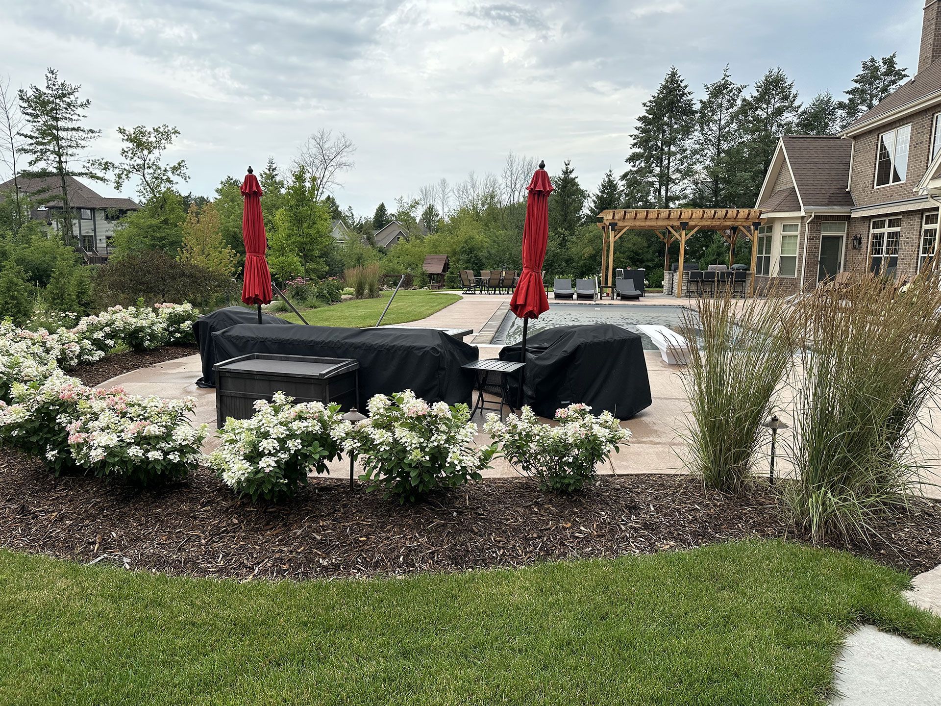 Patio area with covered outdoor furniture and red umbrellas, next to a backyard pool and house with a wooden pergola.