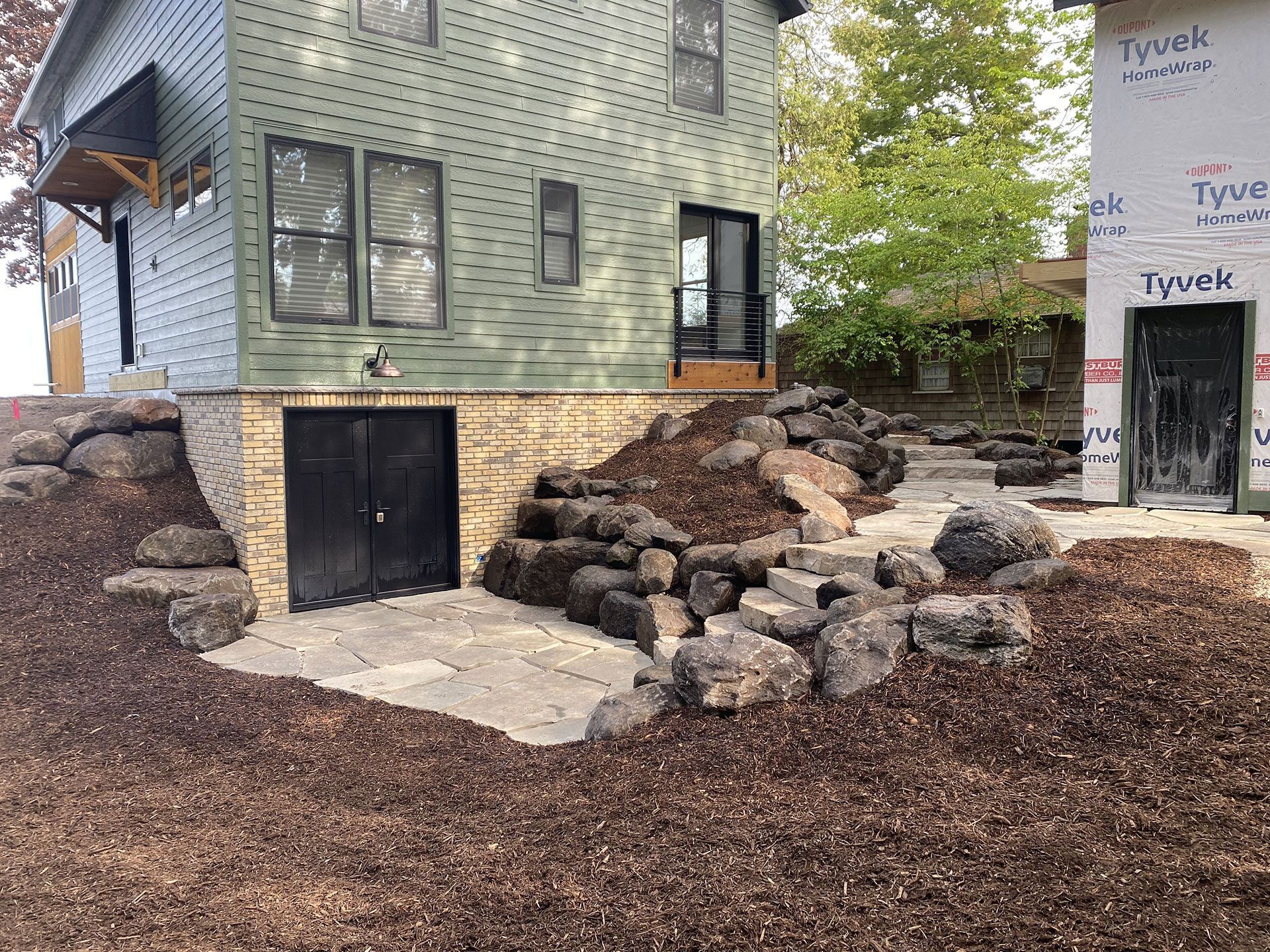 A green two-story house under construction with a stone-retaining wall and patio in a wood-mulched yard.