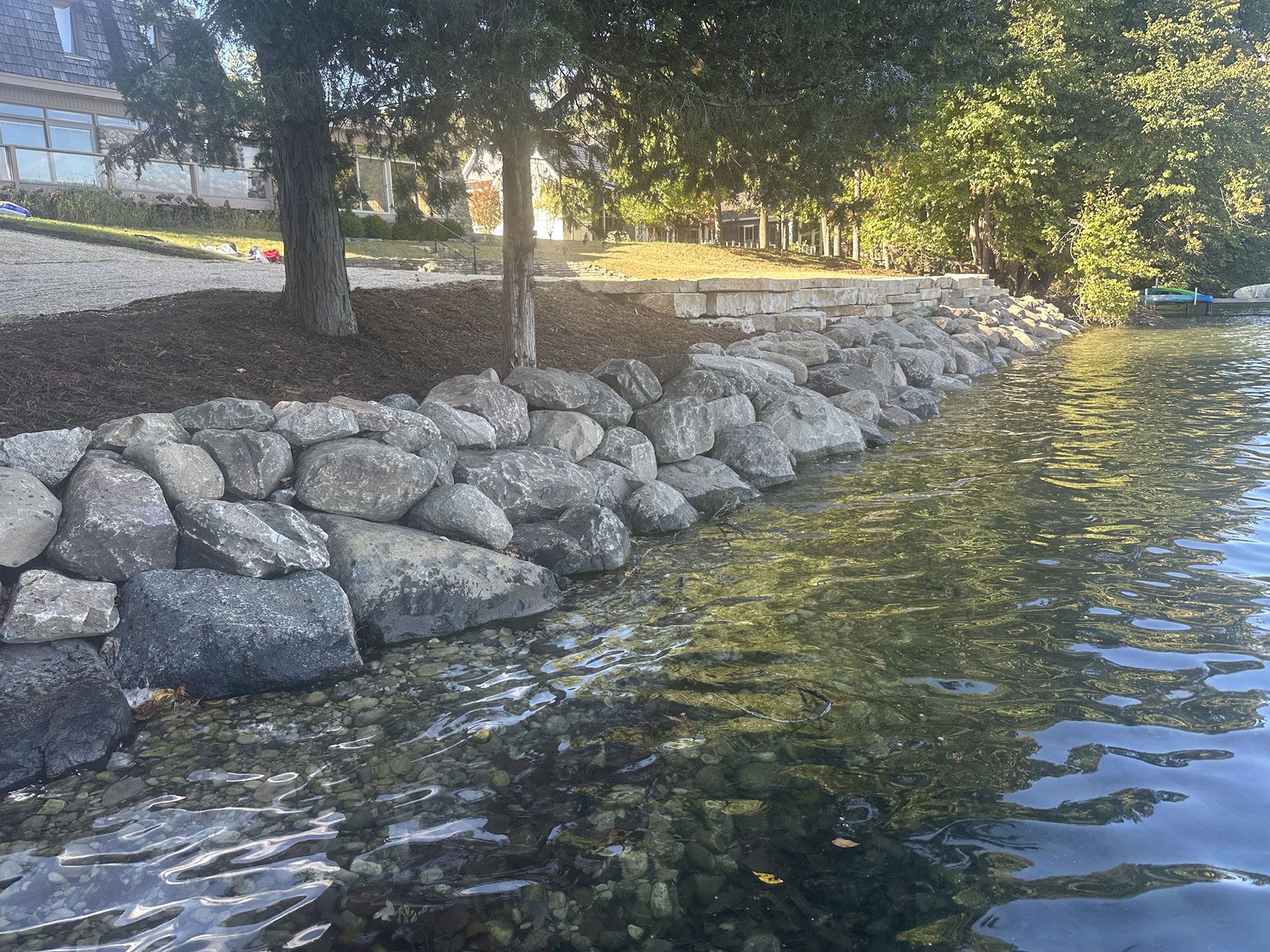 A stone retaining wall bordering a calm lake, with trees and landscaped ground extending behind the shoreline.