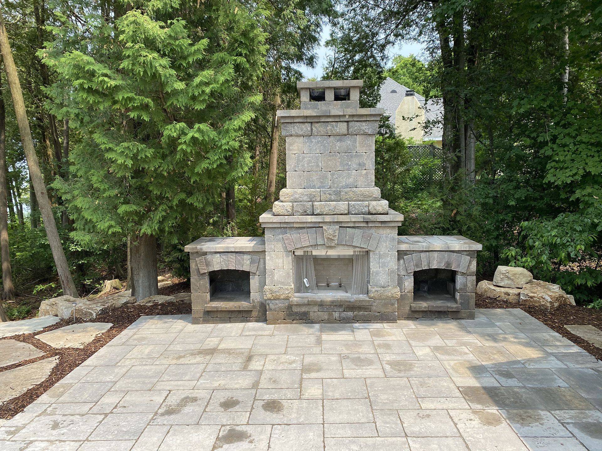 A stone outdoor fireplace with two side compartments, centered on a light-colored paver patio surrounded by trees.