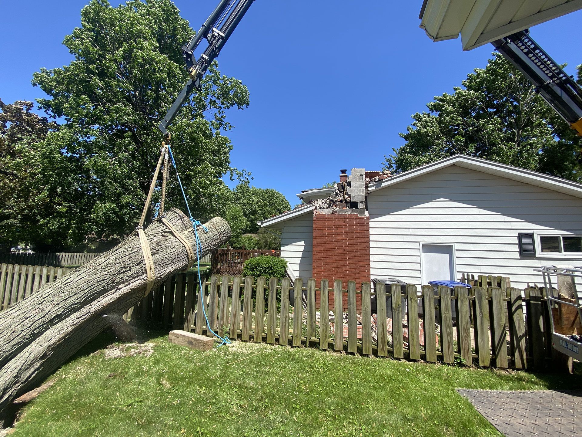 A crane lifts a large tree trunk over a wooden fence in a residential backyard next to a house with a brick chimney.
