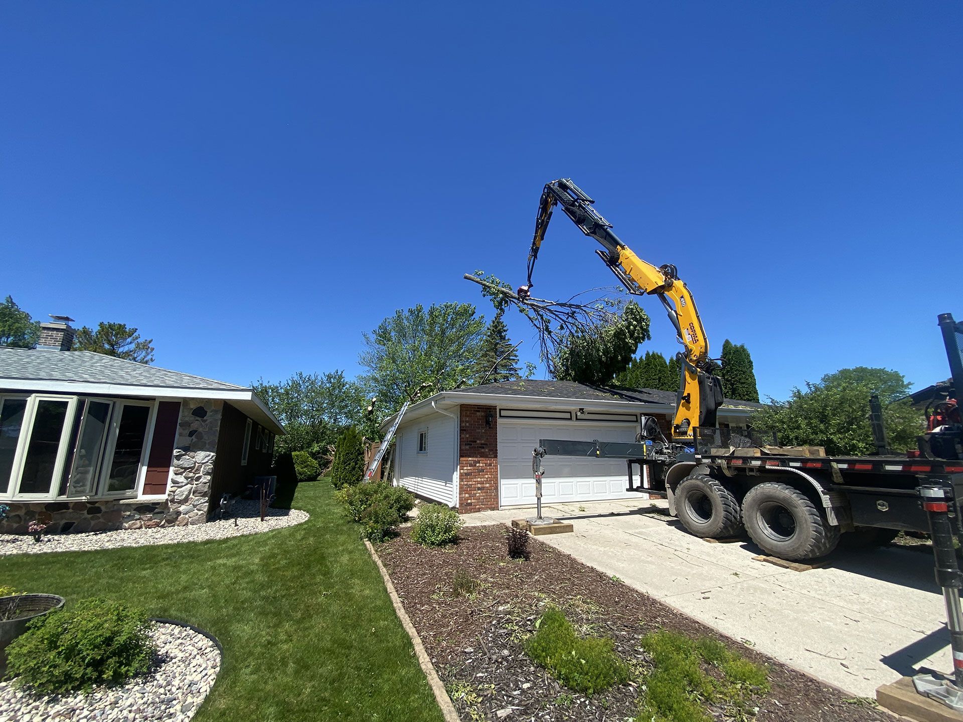 A yellow crane truck parked on a gravel driveway lifts tree branches near the roof of a white garage on a sunny day.