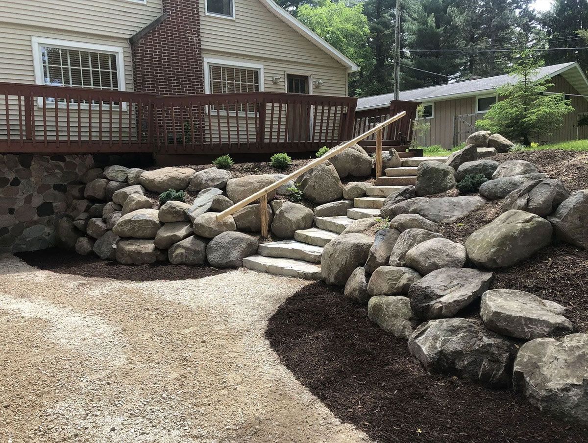 Stone steps with a wooden handrail lead from a gravel path up to a wooden deck in front of a house.