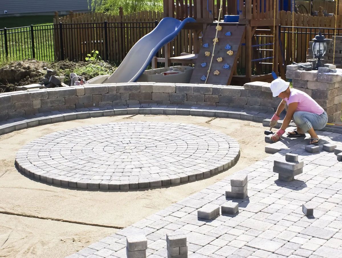 A person wearing a white hat and pink shirt installs pavers to create a circular patio in a backyard.