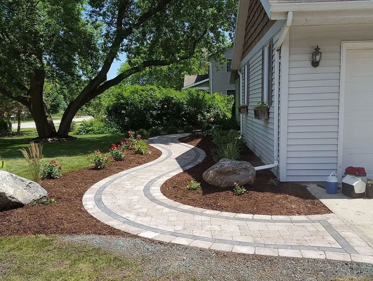 A curved stone paver walkway leads along the side of a light-sided house with mulch beds and decorative landscaping rocks.