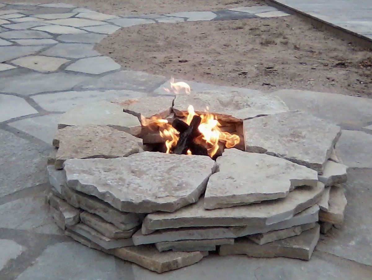 A small, round fire pit built from stacked natural stone slabs sits in the middle of a stone patio with a lit fire inside.