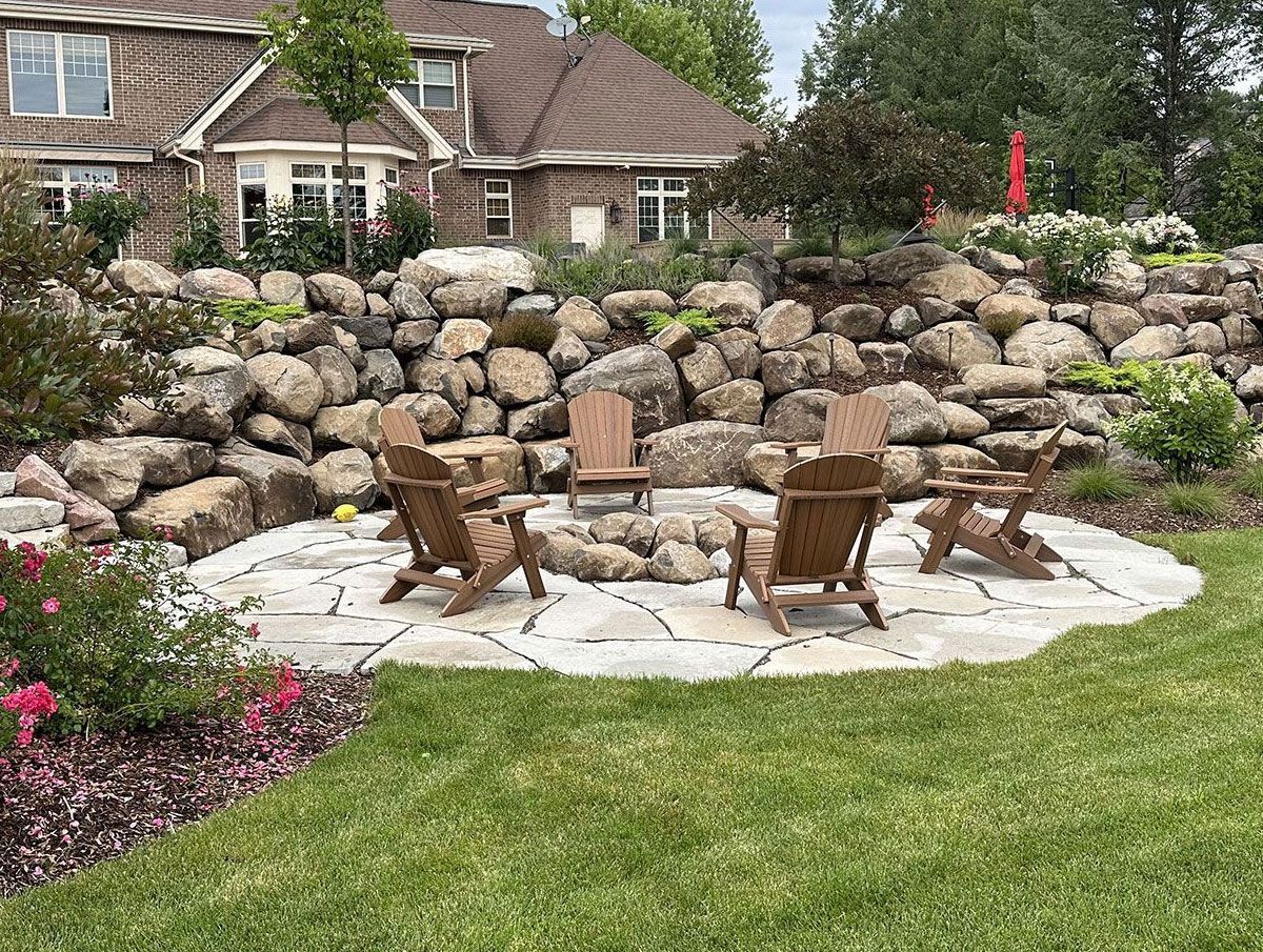 Four wooden chairs sit on a stone patio in a backyard with a large boulder retaining wall behind them.