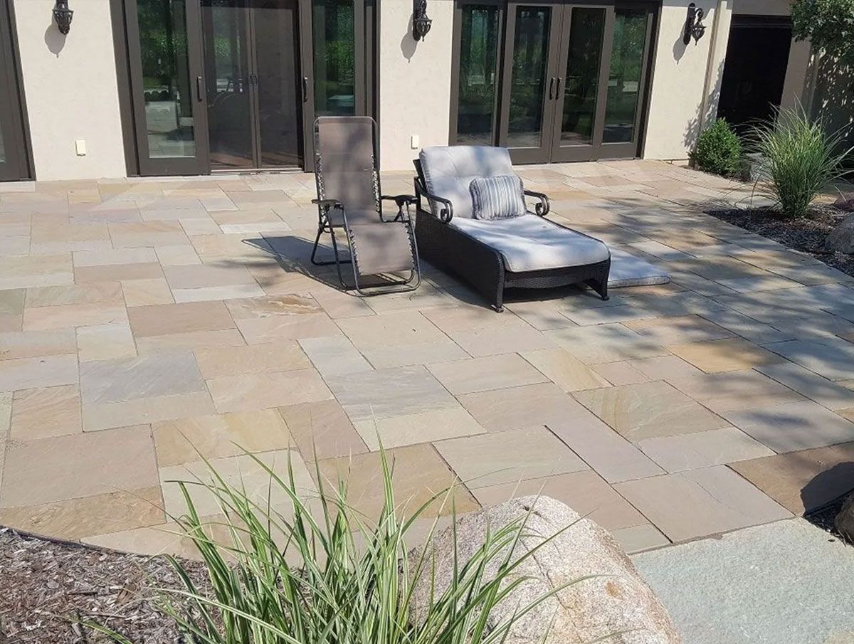 A lounge chair and an upright chair on a large, rectangular-tiled stone patio outside a building with glass doors.