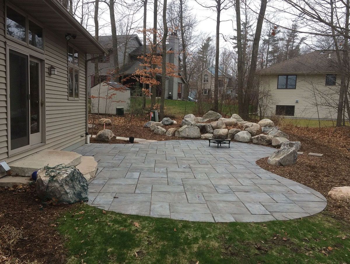 A rectangular gray stone patio sits next to a house, featuring a natural rock garden border in a wooded backyard.
