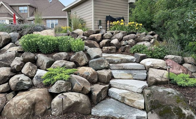 Stone stairs wind through a rock garden with various green shrubs, leading up toward a small shed and residential home.