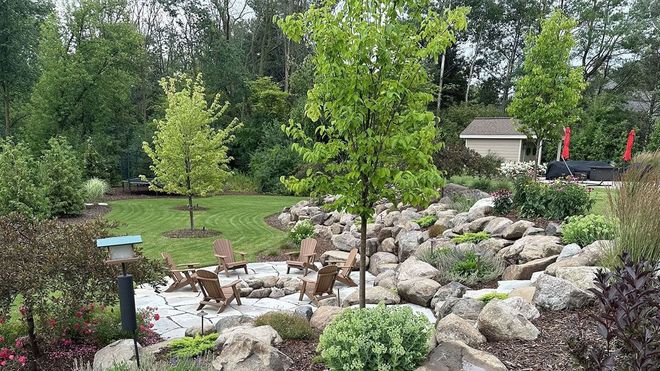 A backyard patio featuring a stone rock garden with Adirondack chairs, trees, and lush green plants.
