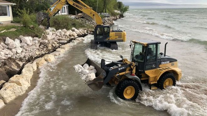 Yellow construction vehicles moving large rocks along a shoreline to build a defensive barrier against incoming waves.