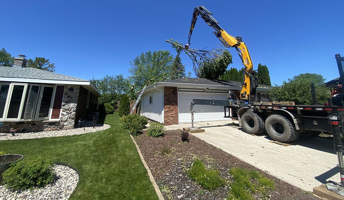 A yellow boom truck crane lifts a large tree over a white garage and house on a sunny day.
