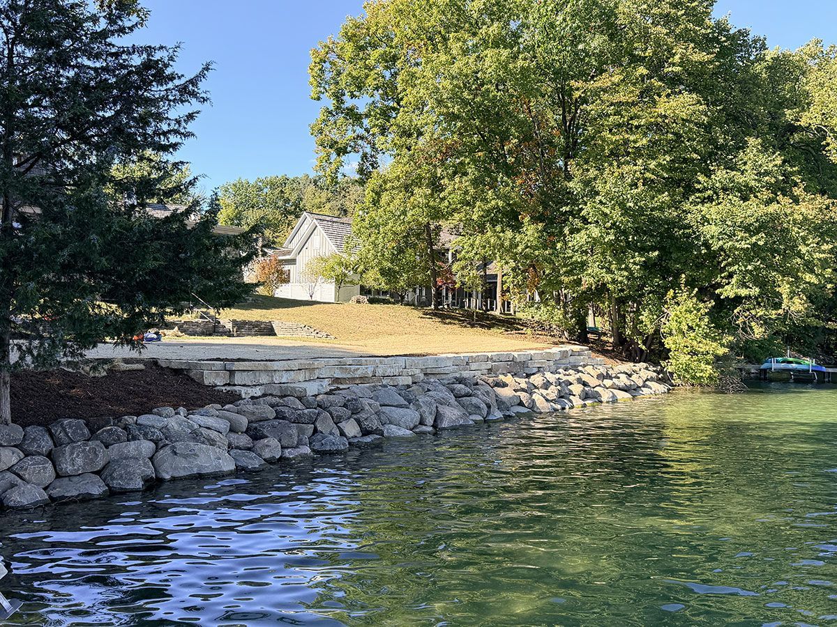 A stone seawall lines the grassy shore of a lake, with a house visible among lush green trees in the background.