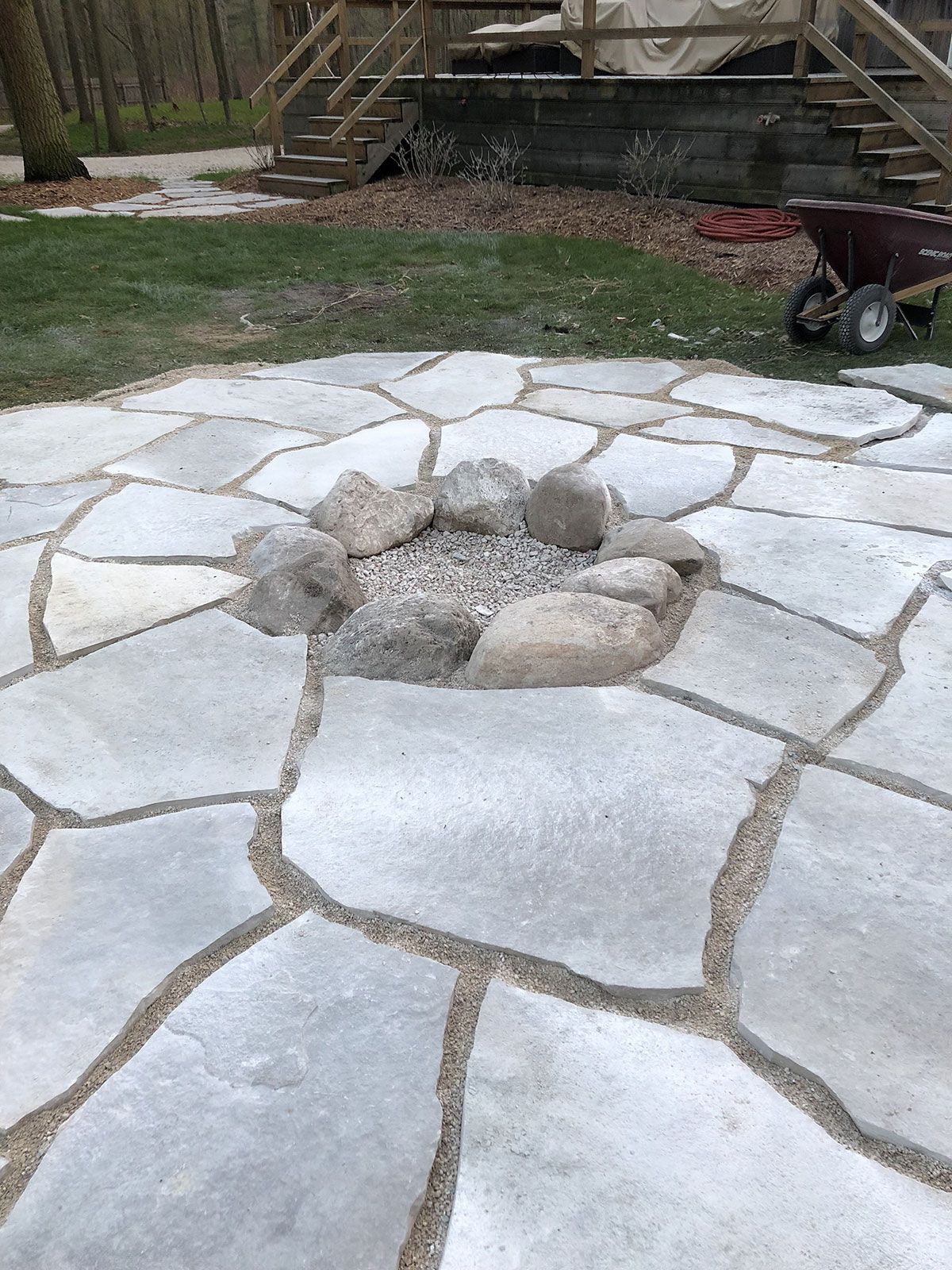 A circular stone patio features a fire pit made of large, grey boulders filled with gravel, set in a backyard.