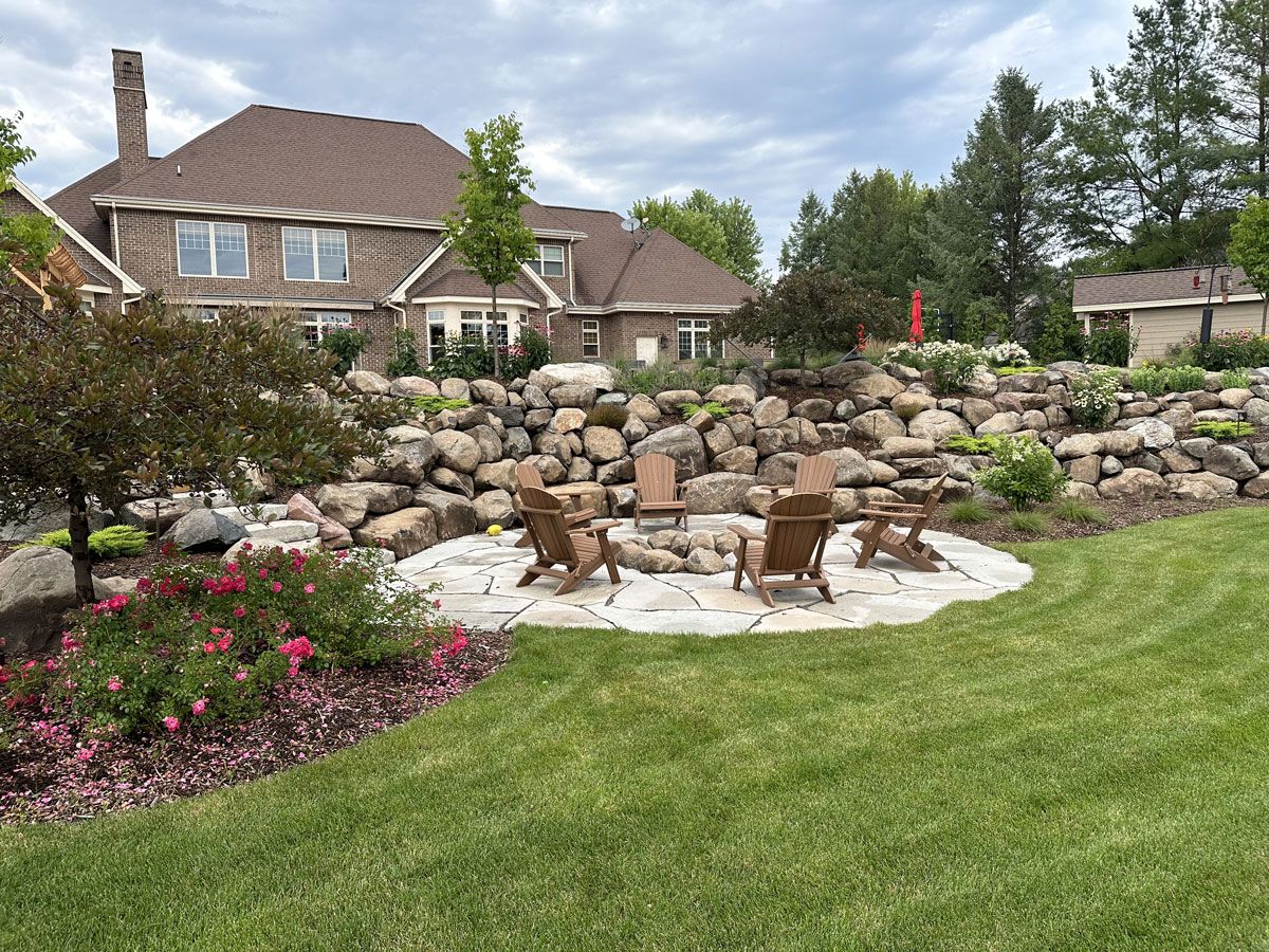 A fire pit area with four chairs sits on a stone patio in front of a large stone retaining wall and a residential house.