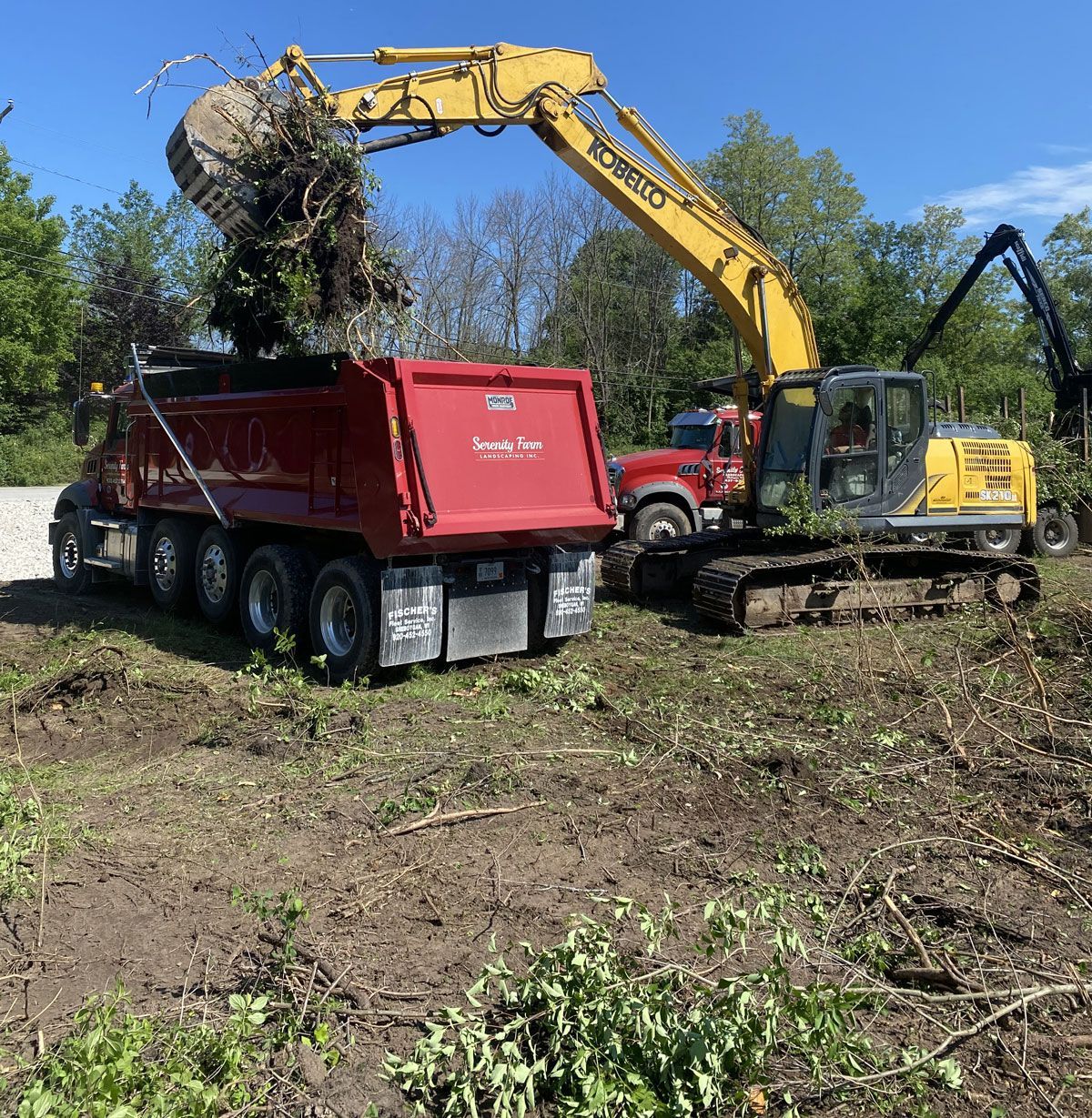 A yellow excavator dumps debris into a red dump truck at a construction site with trees in the background.