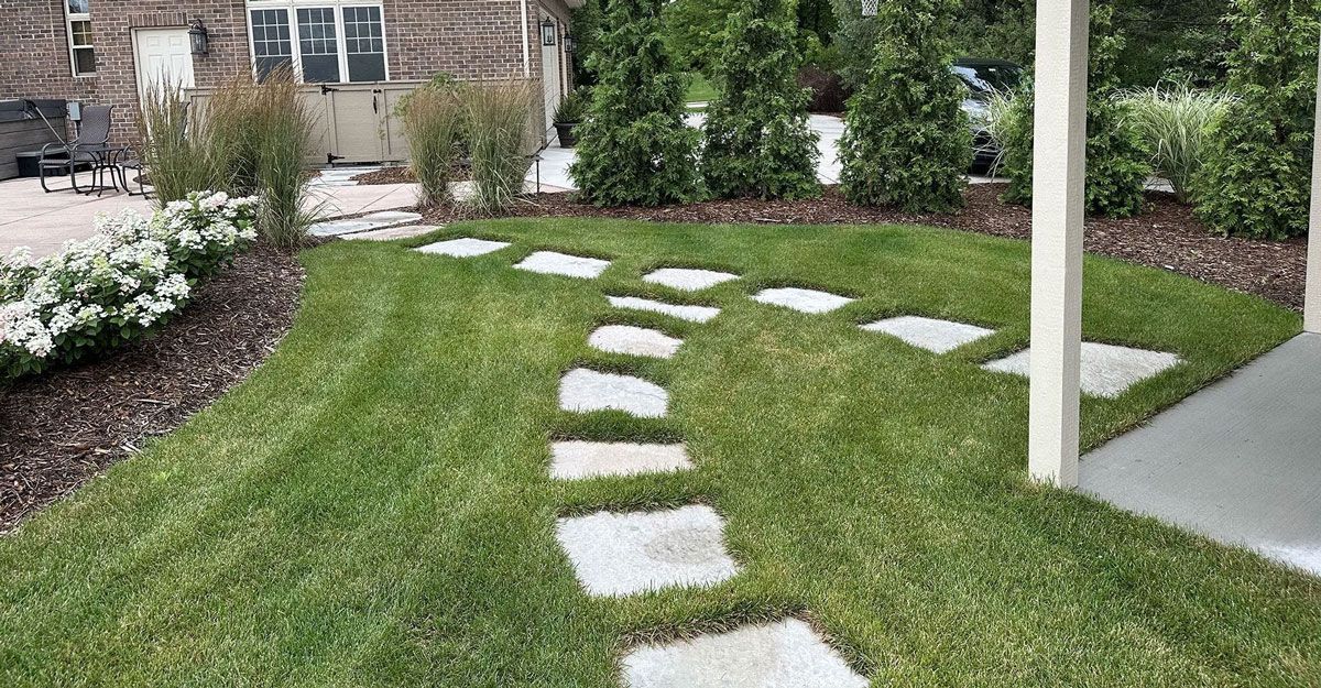A stone path curves through a green lawn beside a house, leading toward a white post and patio edge.