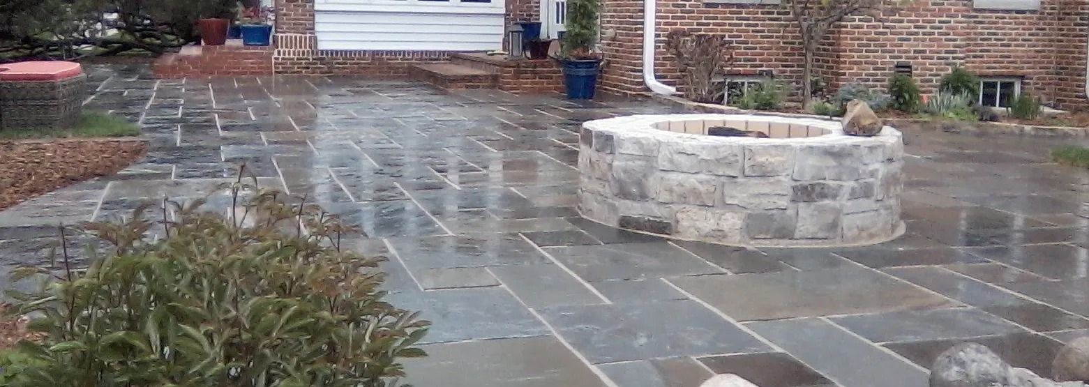 A wet stone patio with a circular stone fire pit in the center, next to a brick house with a white door.