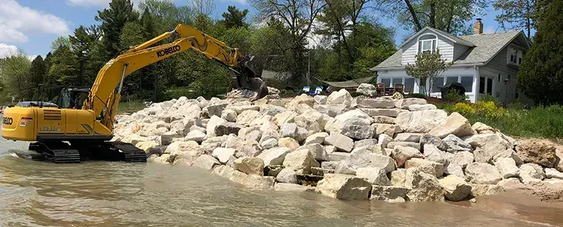 A yellow excavator places large rocks along a shoreline near a house to create a stone retaining wall.
