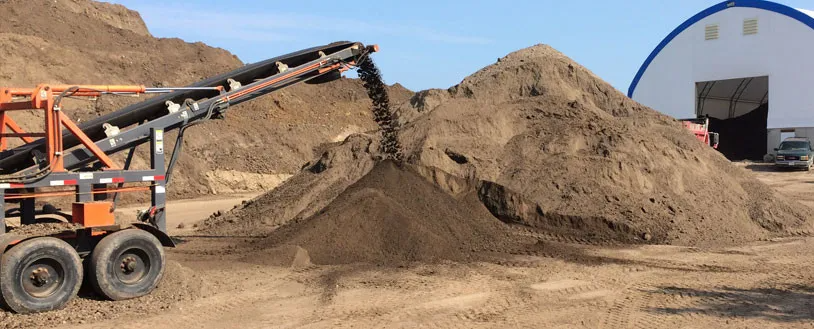 A conveyor belt dumps loose dark soil onto a large mound in front of an industrial storage building.
