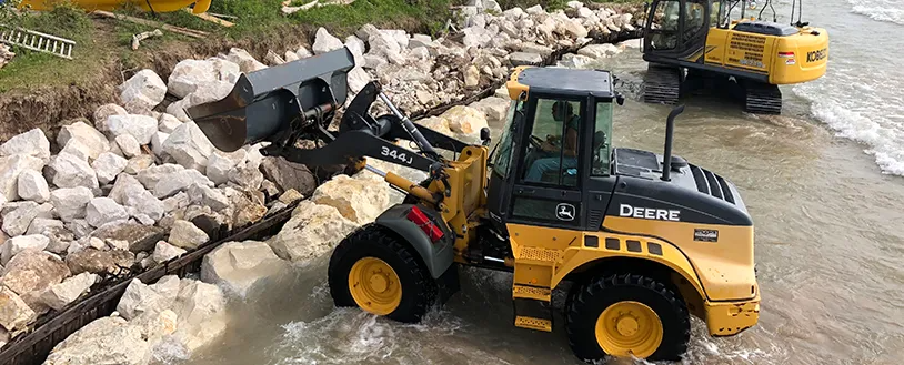 A yellow John Deere wheel loader moves rocks along a shoreline while a yellow excavator works in the water nearby.