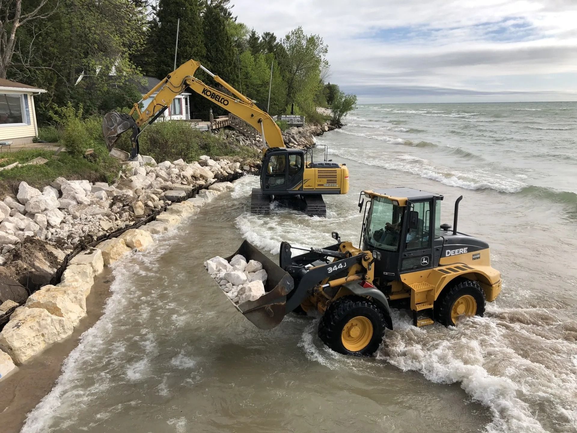An excavator and a wheel loader place large white rocks along a shoreline to prevent erosion by the water.
