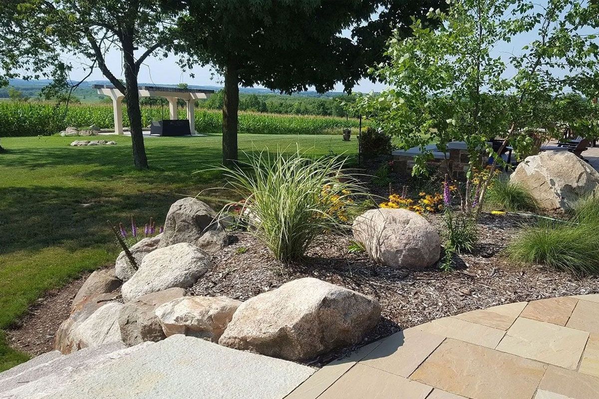 A rock garden with a stone path in the foreground, leading to a grassy yard with trees and a distant field.