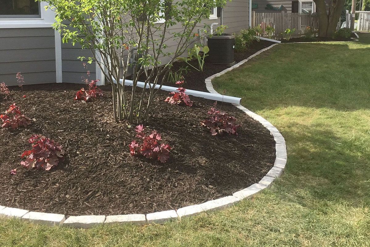 A curved landscape bed with dark mulch, small reddish plants, and a light stone border edging against a green lawn.