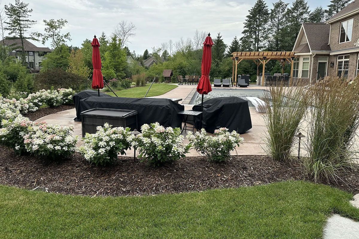 Patio area with covered outdoor furniture, two red umbrellas, white blooming shrubs, and a house in the background.