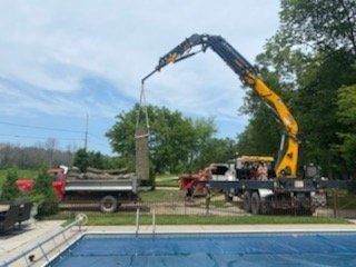 A yellow crane lifts a large log over a swimming pool, moving it toward a red dump truck in a grassy yard.
