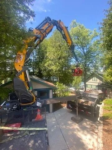 A yellow forestry loader with an extended mechanical arm holds a log above a trailer in a residential yard.