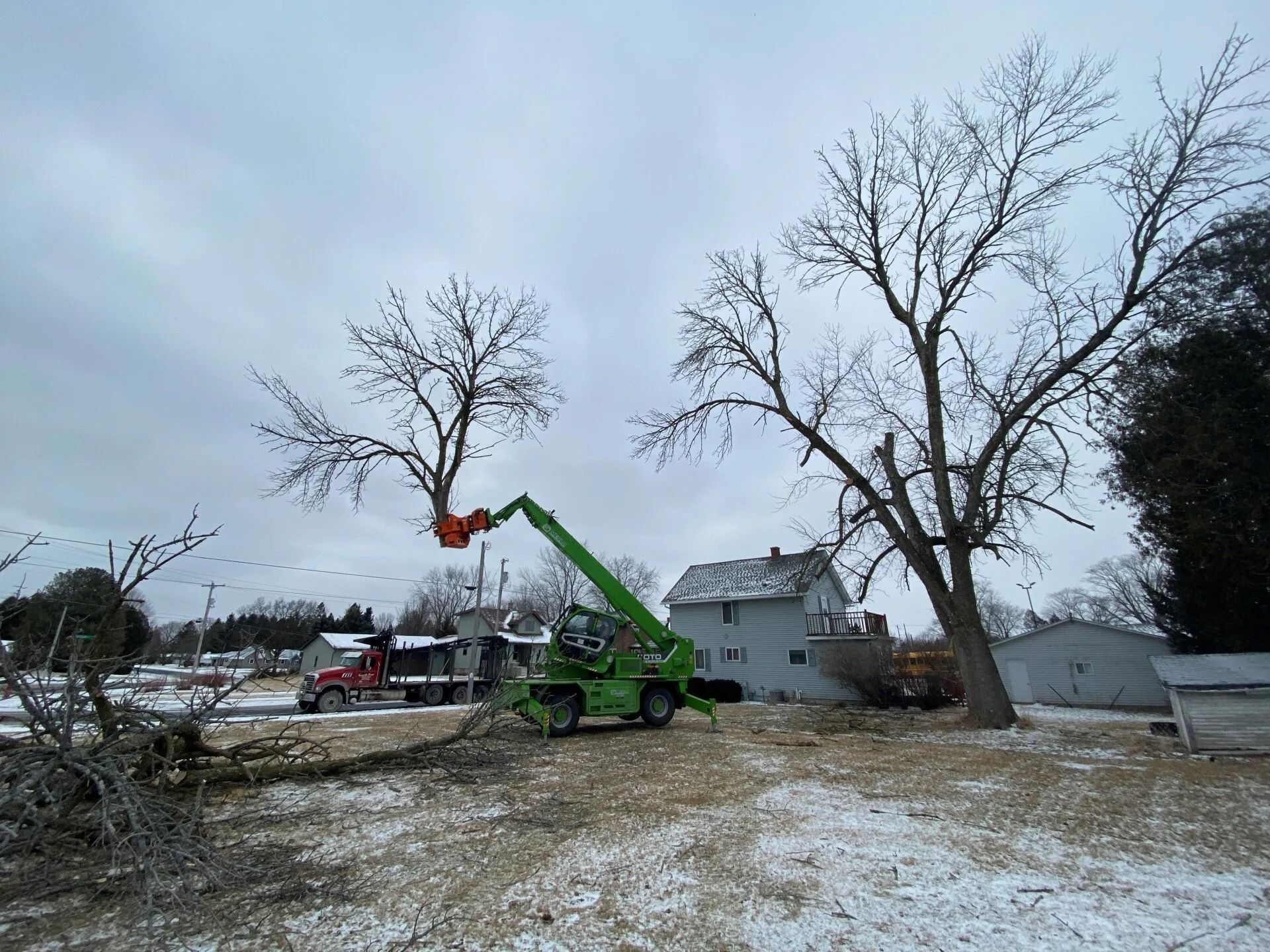 A green tree-cutting crane lifts a severed tree branch in a snow-covered yard near a house and a red truck.
