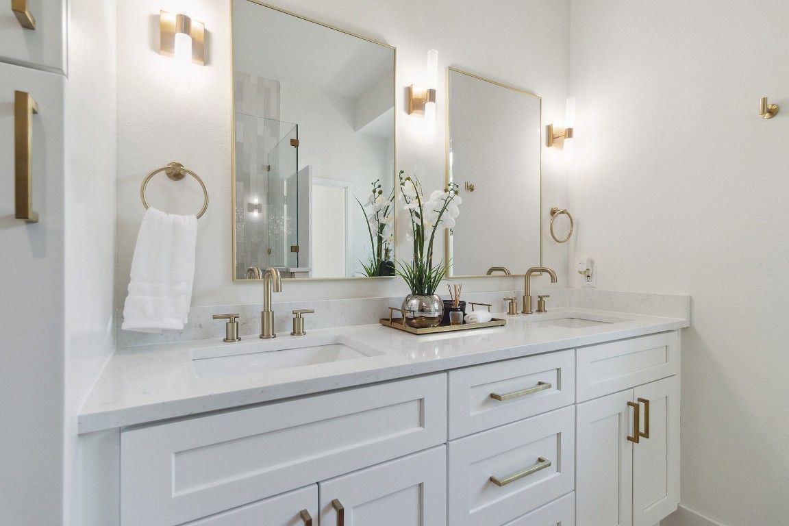 White bathroom with double sinks, gold accents, and mirrors.