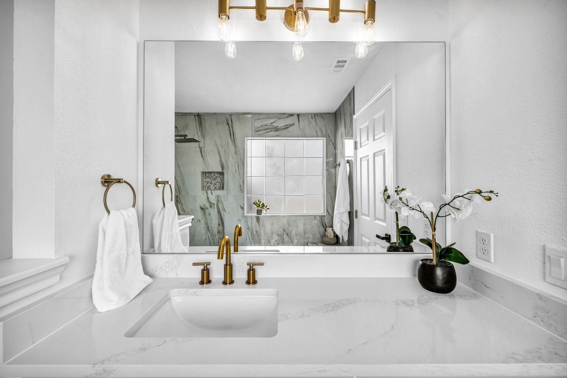 Bathroom with white countertop, gold fixtures, large mirror, and shower in the reflection.
