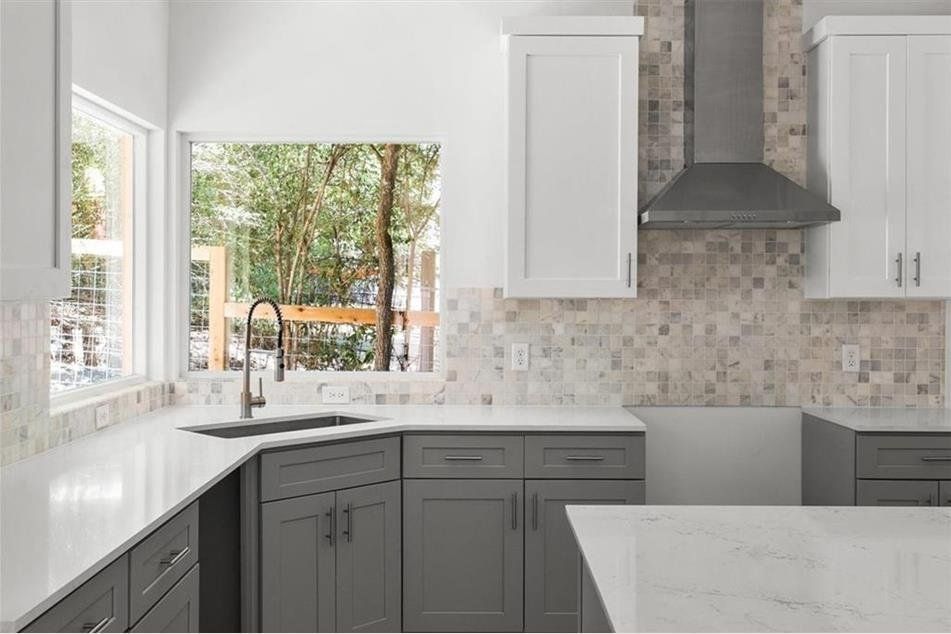 Modern kitchen with gray and white cabinets, stainless steel range hood, and window overlooking trees.