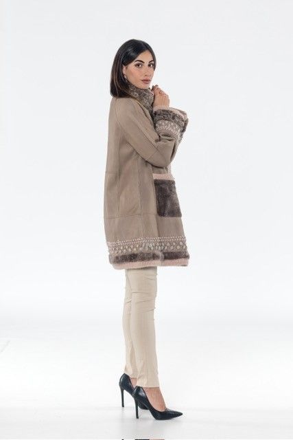 Woman in a tan coat and pants, standing and looking sideways. White backdrop.