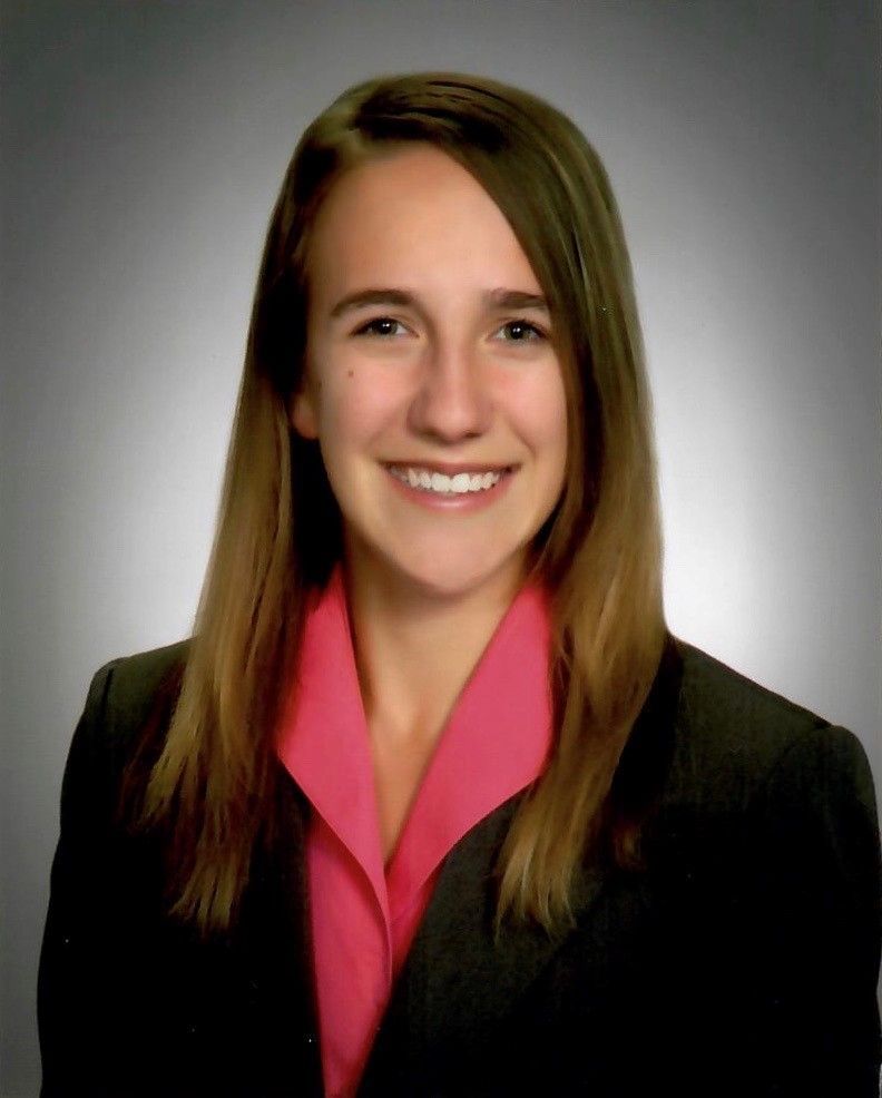 Headshot of a person with shoulder-length brown hair, wearing a dark blazer over a pink collared shirt, smiling on grey.
