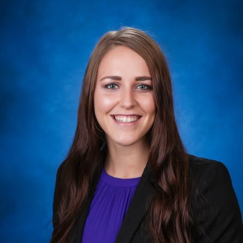 A professional headshot of a smiling person with long brown hair, wearing a purple top and a black blazer, blue background.