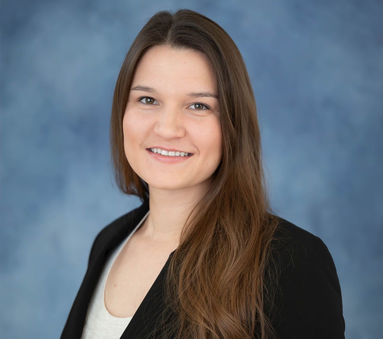 A professional headshot of a person with long brown hair, wearing a black blazer and white top against a blue backdrop.