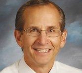 A person with short hair and glasses smiles while wearing a white shirt against a blue, textured background.