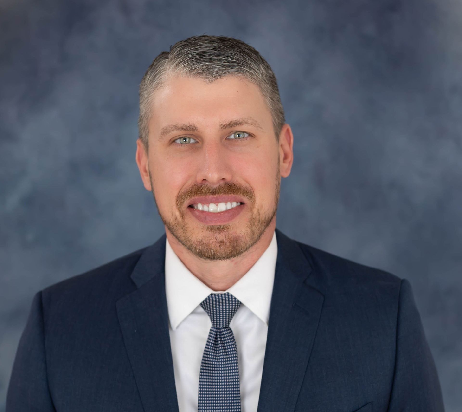 Professional headshot of a smiling person wearing a dark blue suit, white shirt, and patterned tie against a grey background.