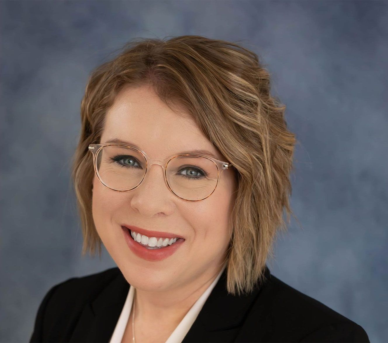 A smiling person with shoulder-length blonde hair and glasses, wearing a dark blazer over a white shirt against a blue wall.