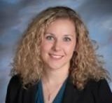 A person with curly blonde hair, wearing a dark blazer over a teal shirt, smiles against a blue, textured background.