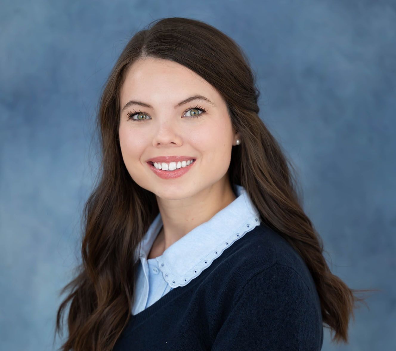 A person with long brown hair smiling against a textured blue backdrop, wearing a dark sweater over a collared shirt.